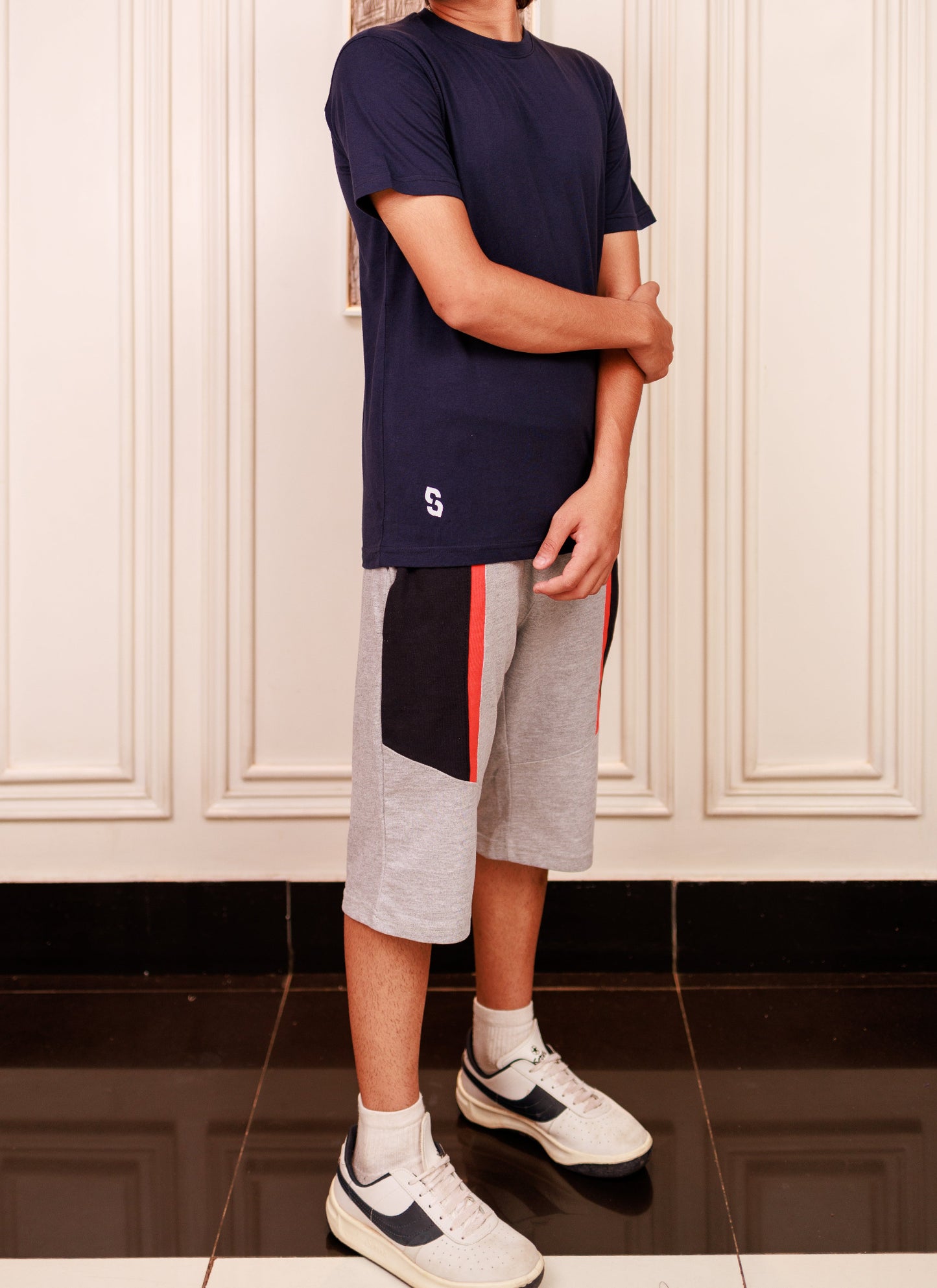 Person wearing a navy t-shirt and gray shorts with a red stripe, standing in a room with white paneled walls.