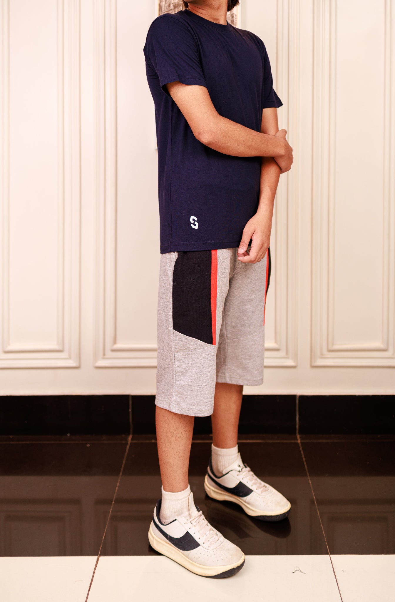 Person wearing a navy t-shirt, gray shorts with red and black stripes, and white sneakers in a room.