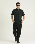 A man standing against a white background wearing a short-sleeved black shirt with a collar, black pants, and sunglasses.