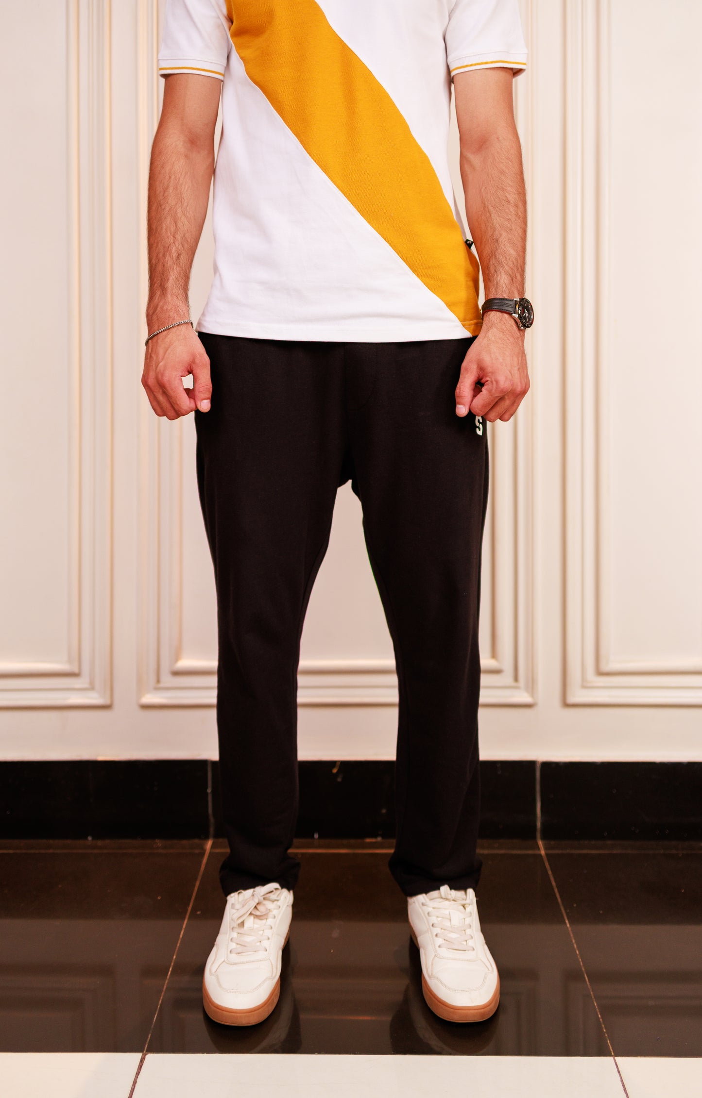Person wearing a white t-shirt with a yellow stripe, black pants, and white sneakers in a room.