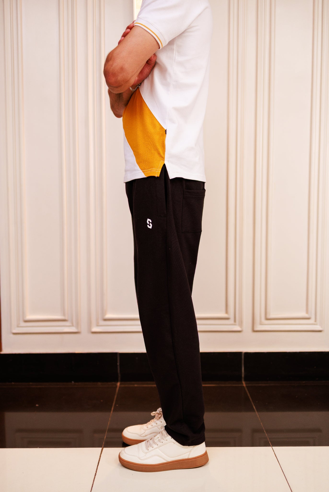 Person wearing a white and yellow top with black pants in a room with white paneled walls.