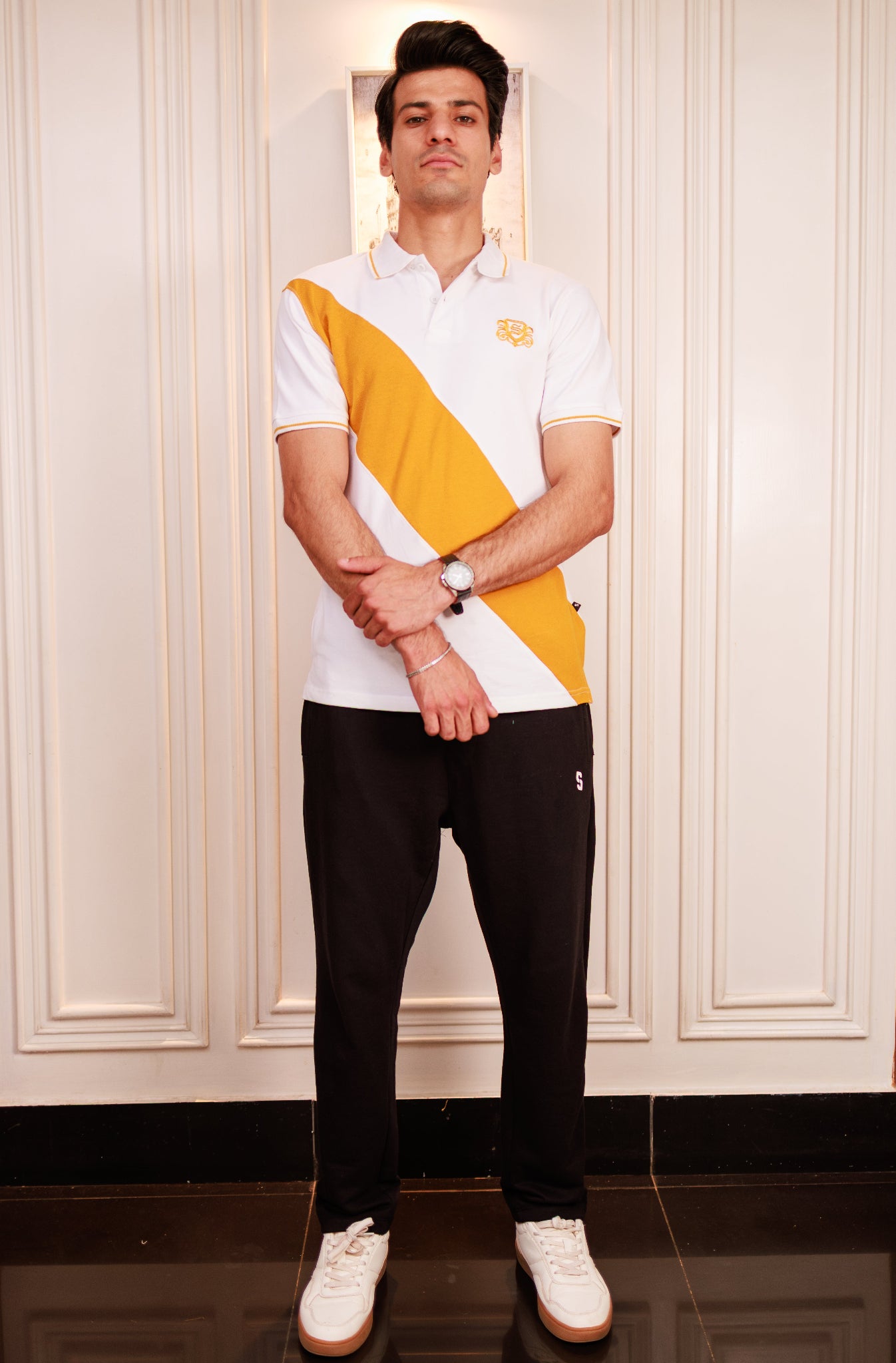 Man wearing a white and yellow striped polo shirt with black pants, standing against a white paneled wall.