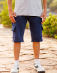 Person wearing blue cargo shorts and white sneakers on a stone path with greenery in the background