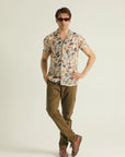 A man wearing sunglasses, a short-sleeved geometric patterned shirt with a resort collar, and brown trousers.
