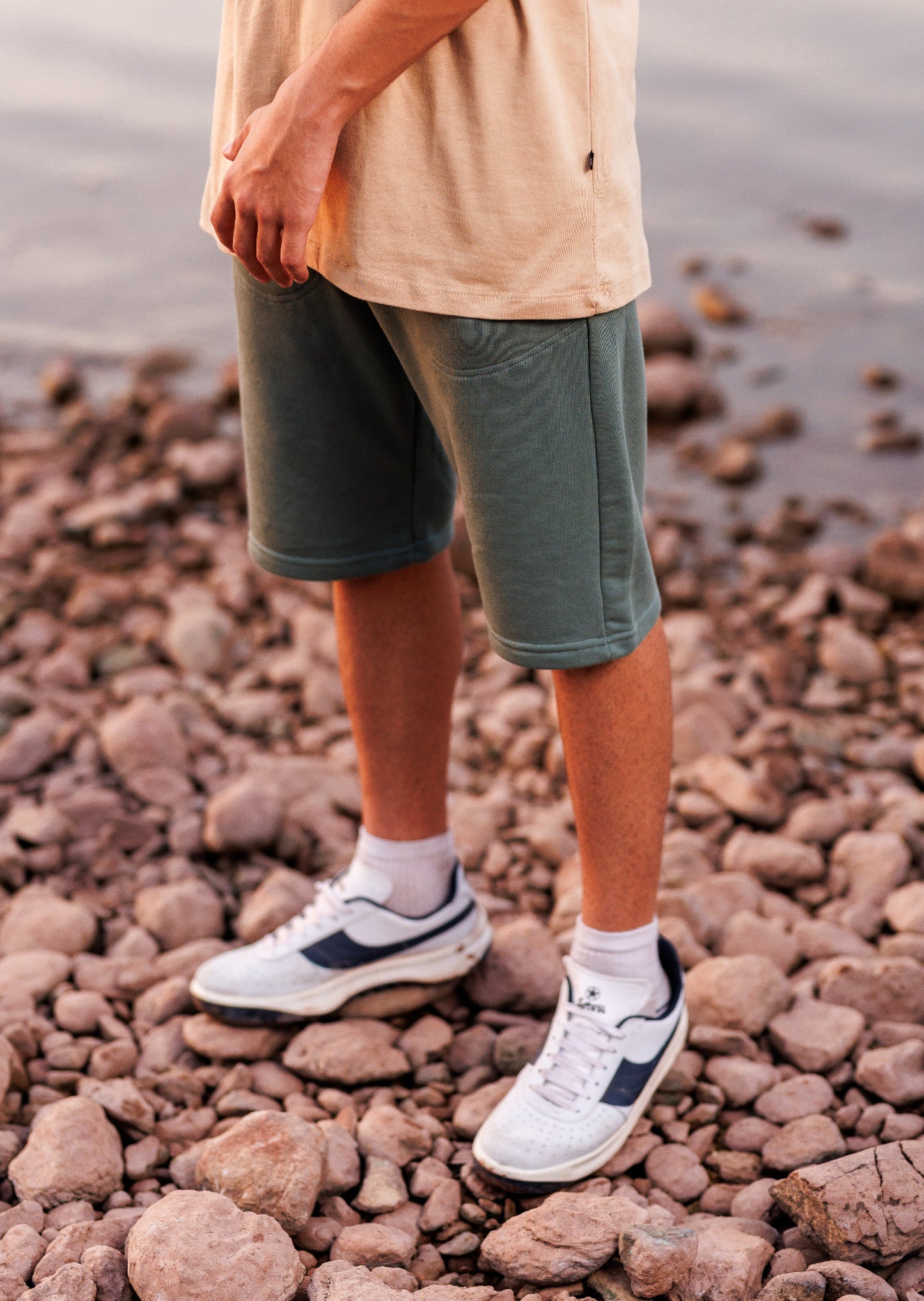 Person wearing green shorts and white sneakers standing on a rocky beach.