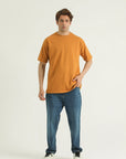 A man wearing a yellow loose fit t-shirt with a rib-trimmed neckline, dropped shoulders, and a straight-cut hem, paired with blue jeans and white sneakers.