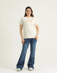 A woman standing against a white background wearing a white round neck t-shirt with gold metallic print and blue flare jeans.