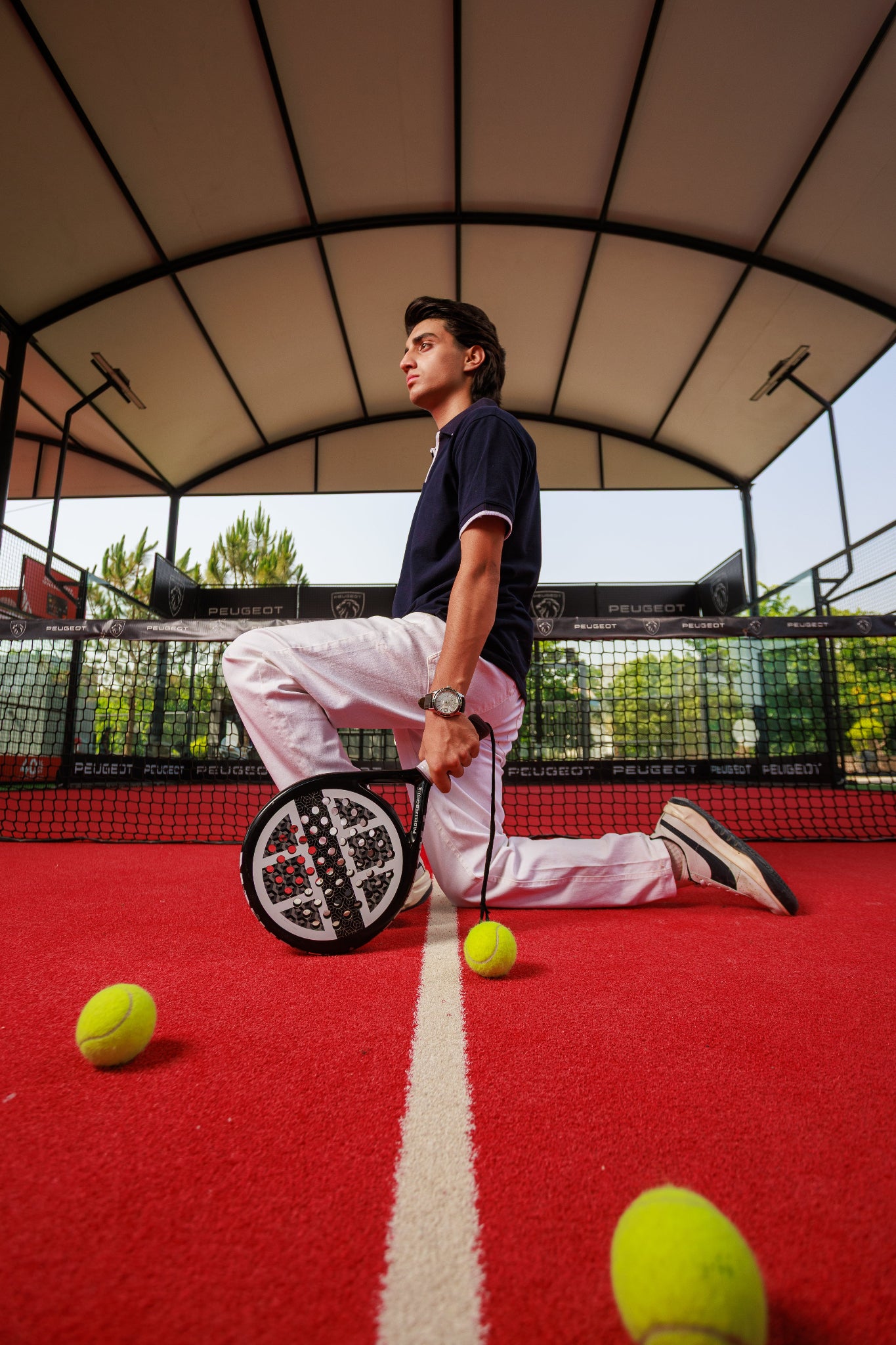 Person on a tennis court with a racquet and balls, under an open roof.