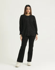 A woman standing against a white background wearing a black long-sleeve top with lace detailing at the neck and front, and black pants.