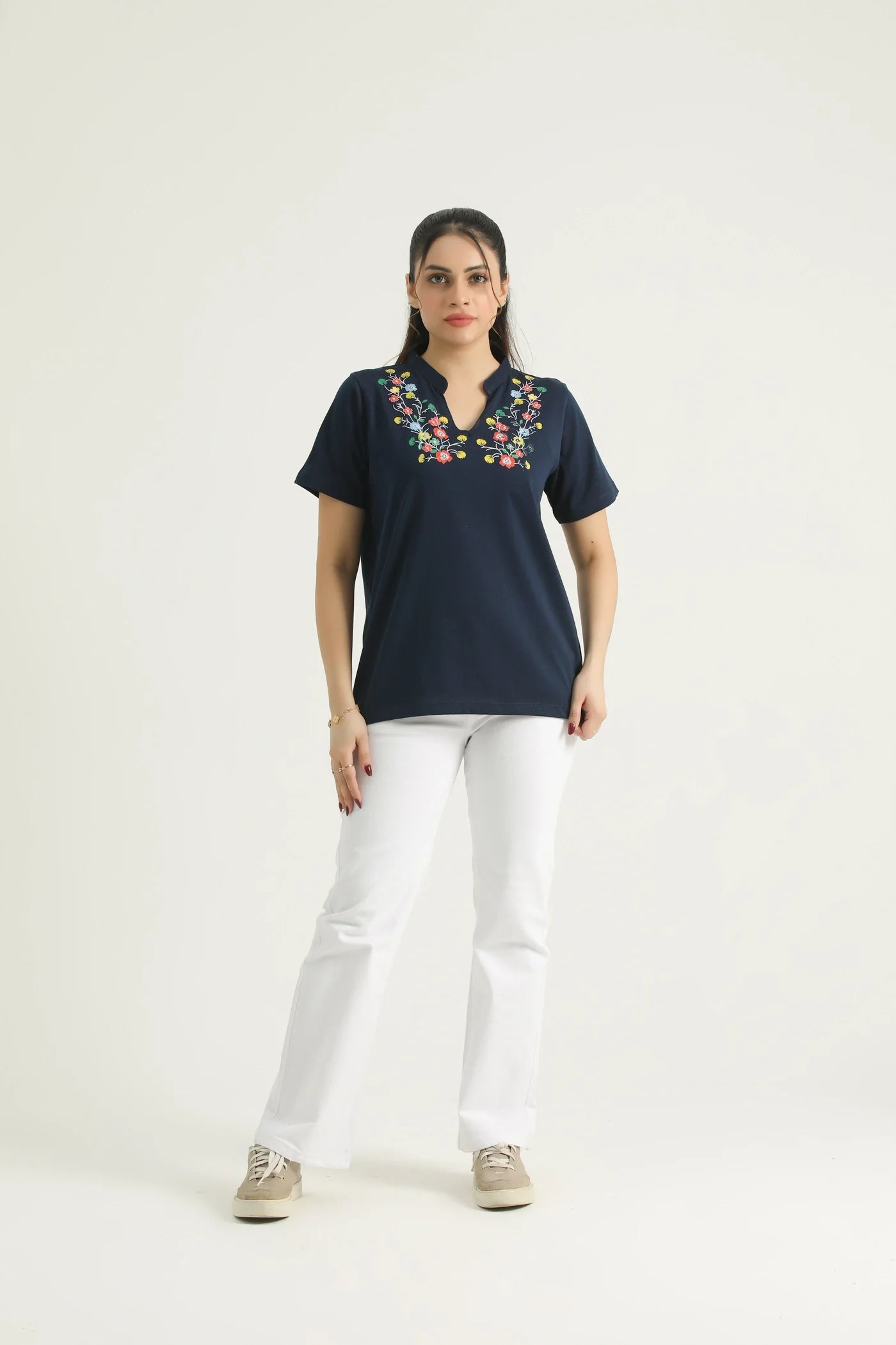 A woman standing against a white background wearing a navy blue knit top with a floral print and a V-neckline, paired with white pants and beige shoes.