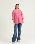 A woman standing against a white background wearing a blush pink top with a scalloped V-neck and three-quarter straight sleeves, paired with blue jeans and white shoes.