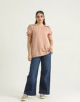 A woman standing against a white background wearing a pink top with round neckline, pleats at shoulders, and elasticated half puffy sleeves, paired with blue jeans.