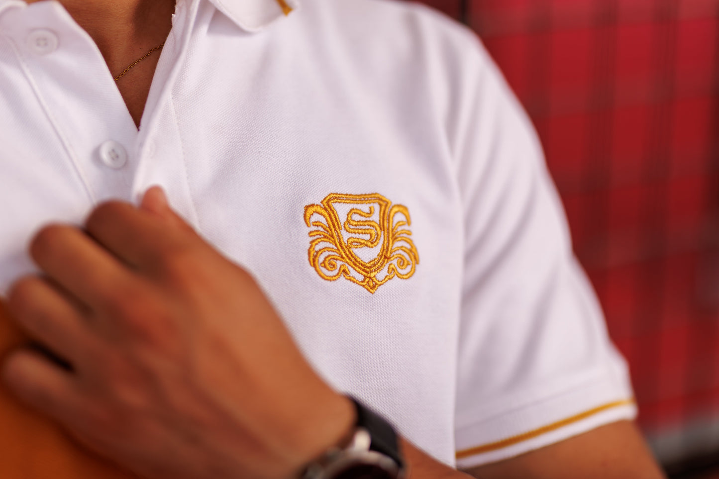 White polo shirt with gold emblem worn by a person, blurred red background