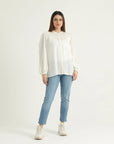 A woman wearing a white button-down shirt with gathers at the yoke and shoulder edges, elasticated pleated long sleeves, paired with blue jeans and white shoes.