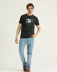 A man wearing a black round-neck t-shirt with a graphic print in front and light blue jeans. He is also wearing brown shoes and has a watch on his left wrist.
