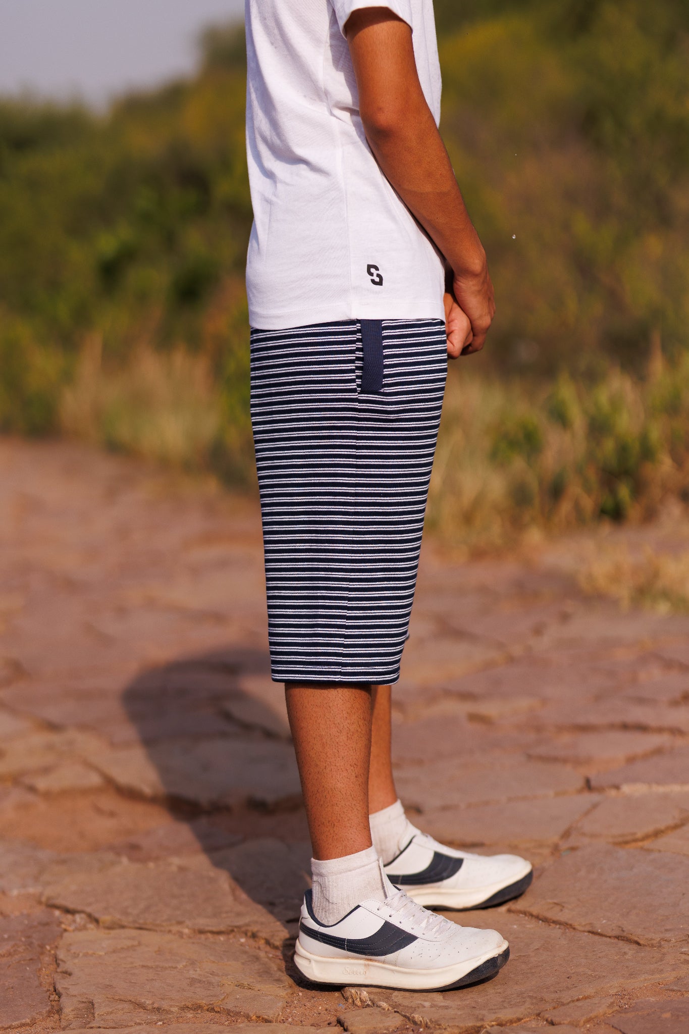 Person wearing a white t-shirt, blue striped shorts, and white sneakers on a stone path with grass in the background.