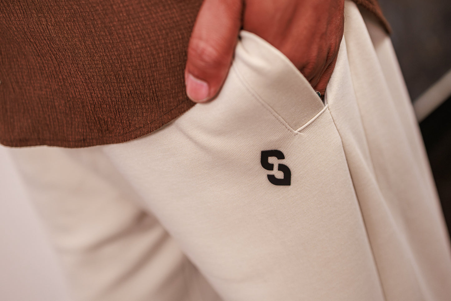 Close-up of beige pants with a logo on the pocket, worn by a person in a brown sweater.
