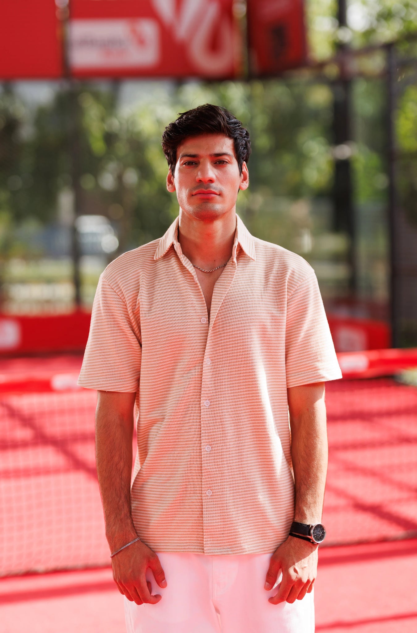 Man standing on a tennis court wearing a light pink shirt and white pants.