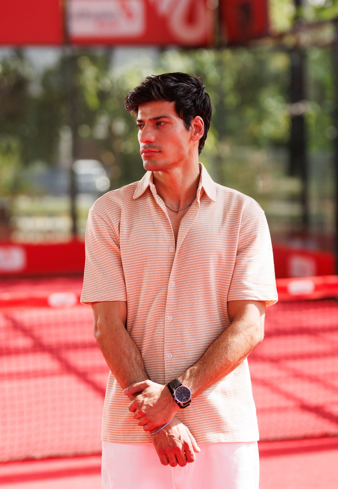 Man standing on a tennis court wearing a light pink shirt and white pants.