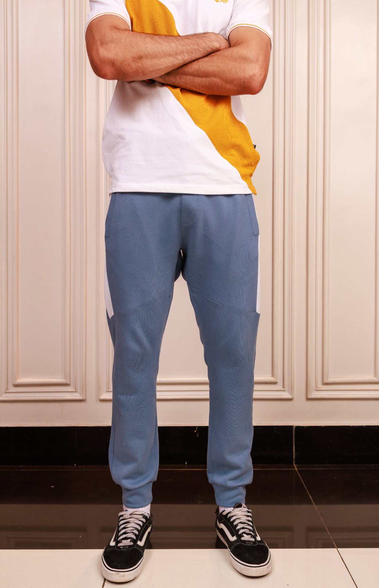 Person wearing a white and yellow shirt with blue pants standing against a beige wall.