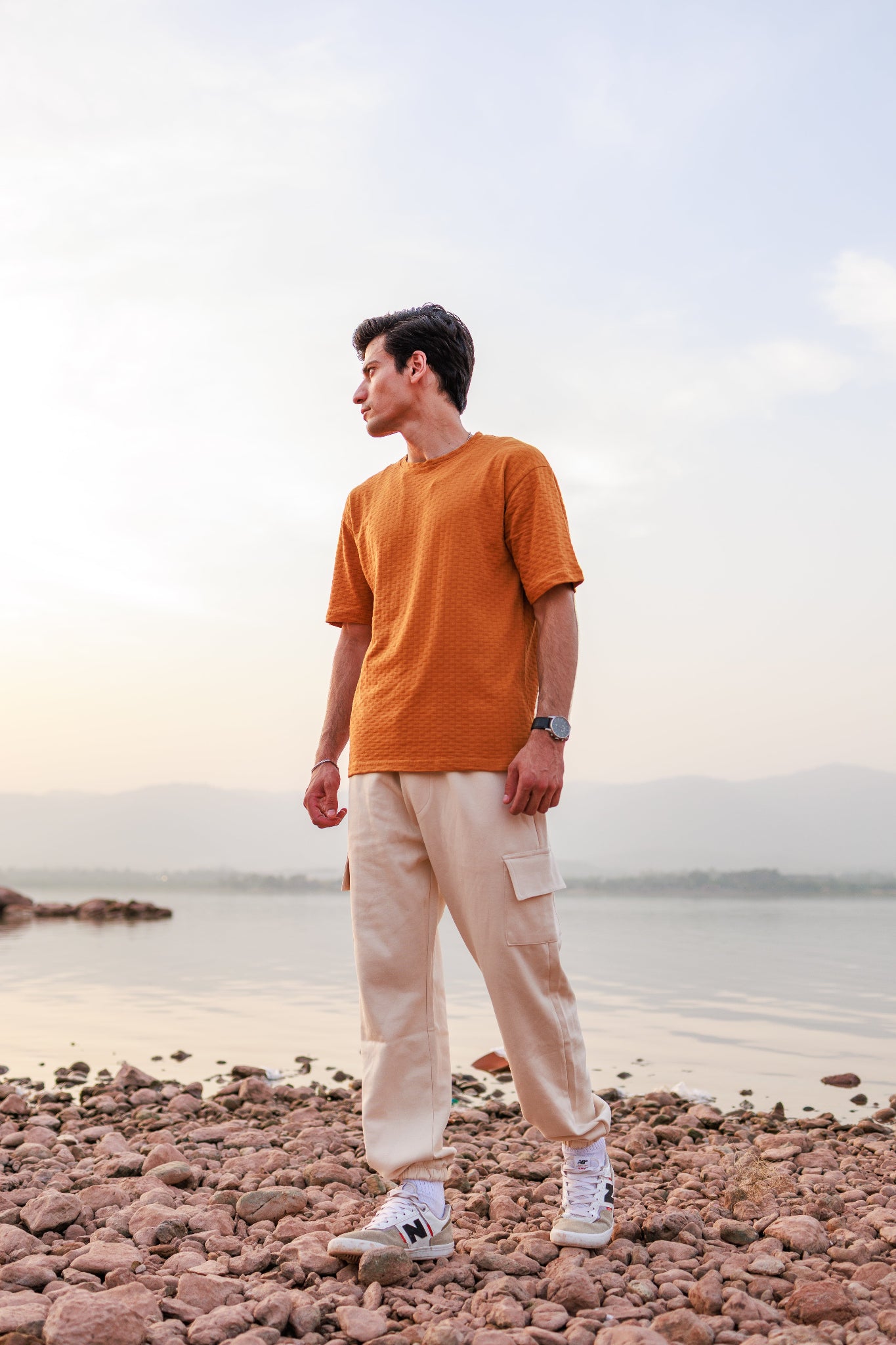 Man standing on a rocky beach wearing an orange shirt and beige pants.