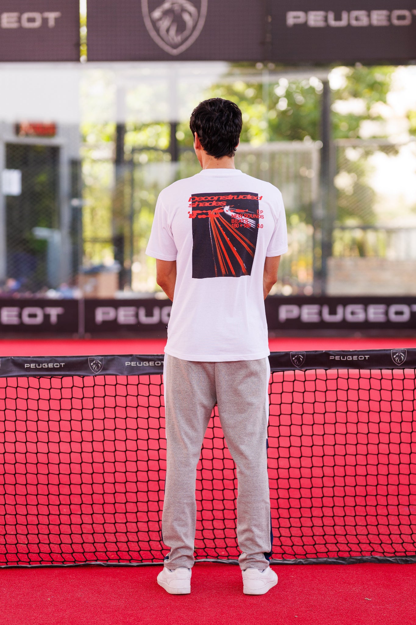 Person standing on a red tennis court with a Peugeot banner in the background