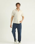 A man standing against a white background wearing a beige short-sleeved button-down shirt with a Cuban collar and blue jeans.