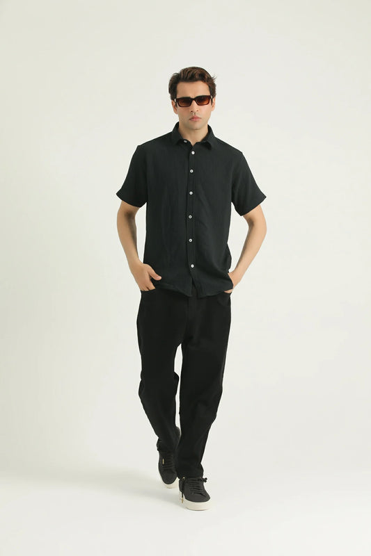 A man standing against a white background wearing a short-sleeved black shirt with a collar, black pants, and sunglasses.