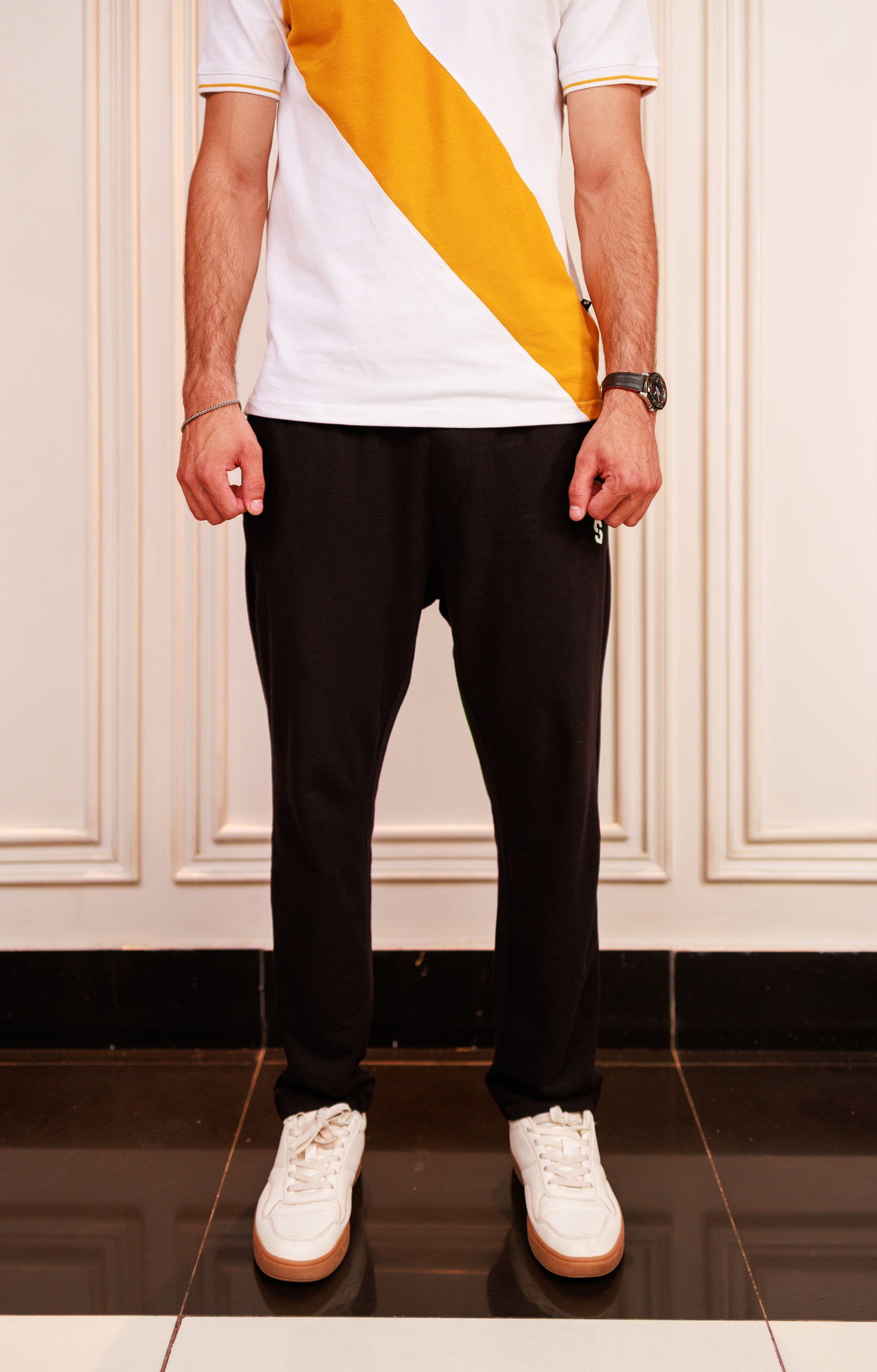 Person wearing a white t-shirt with a yellow stripe, black pants, and white sneakers in a room.