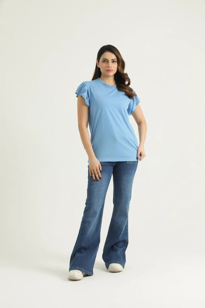 A woman standing against a white background wearing a powder blue frill knit top with frill cap sleeves and round neck, paired with blue jeans and white shoes.