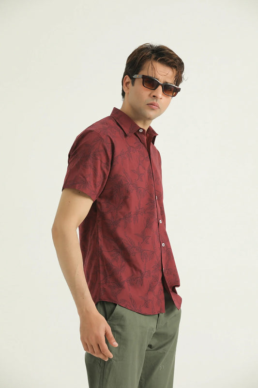 A man wearing a short-sleeved red and black patterned shirt with a collar, sunglasses, and light green trousers.