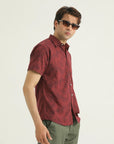 A man wearing a short-sleeved red and black patterned shirt with a collar, sunglasses, and light green trousers.