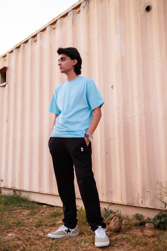 Person wearing a light blue t-shirt and black pants standing against a beige wall.