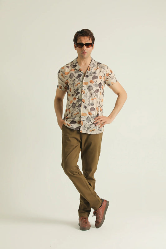 A man wearing sunglasses, a short-sleeved geometric patterned shirt with a resort collar, and brown trousers.
