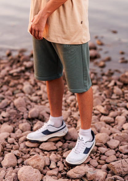 Person wearing green shorts and white sneakers standing on a rocky beach.