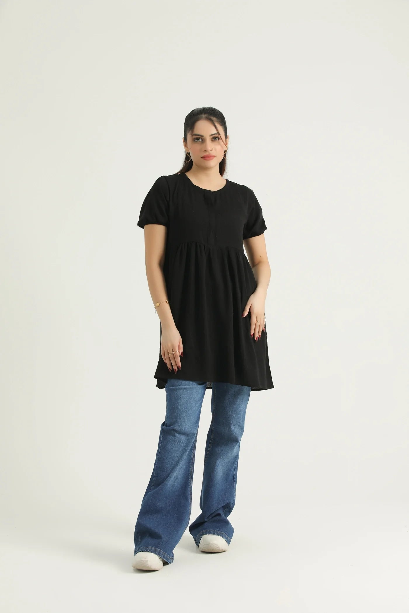 A woman standing, wearing a mid-length black top with short sleeves and a round neckline, paired with blue jeans.