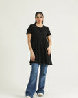 A woman standing, wearing a mid-length black top with short sleeves and a round neckline, paired with blue jeans.
