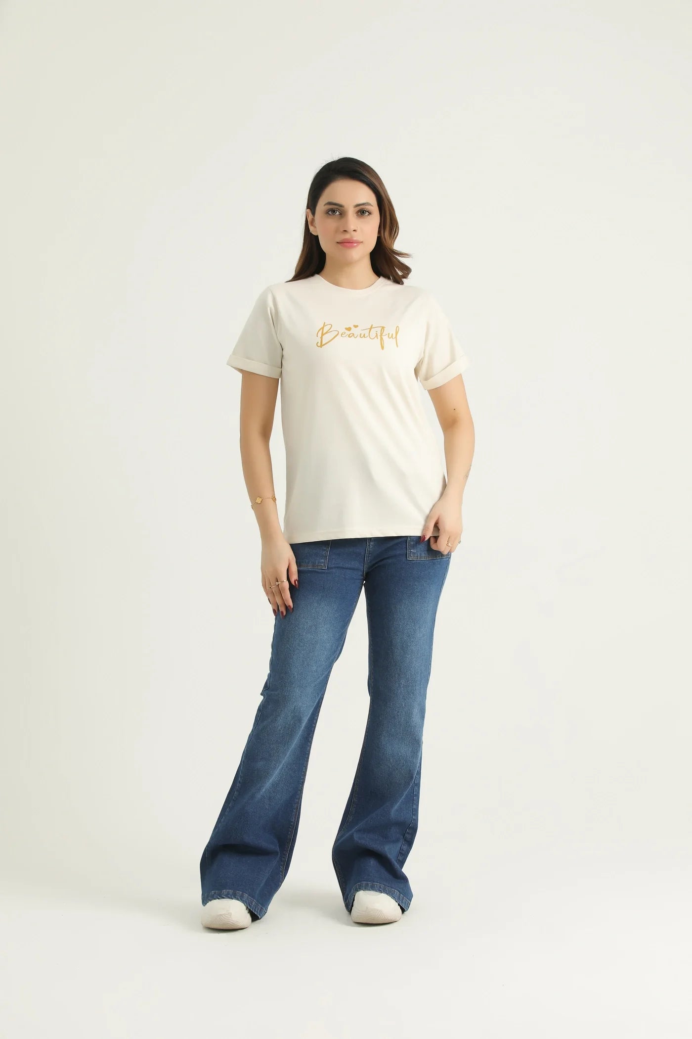 A woman standing against a white background wearing a white round neck t-shirt with gold metallic print and blue flare jeans.