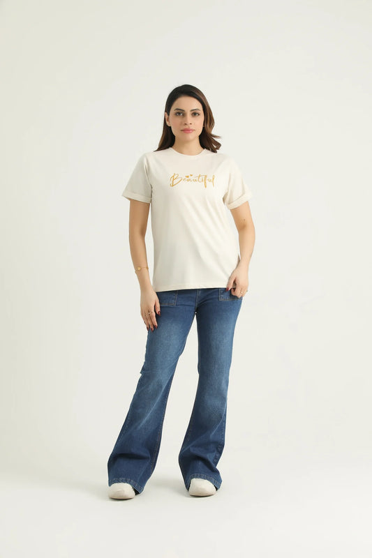 A woman standing against a white background wearing a white round neck t-shirt with gold metallic print and blue flare jeans.