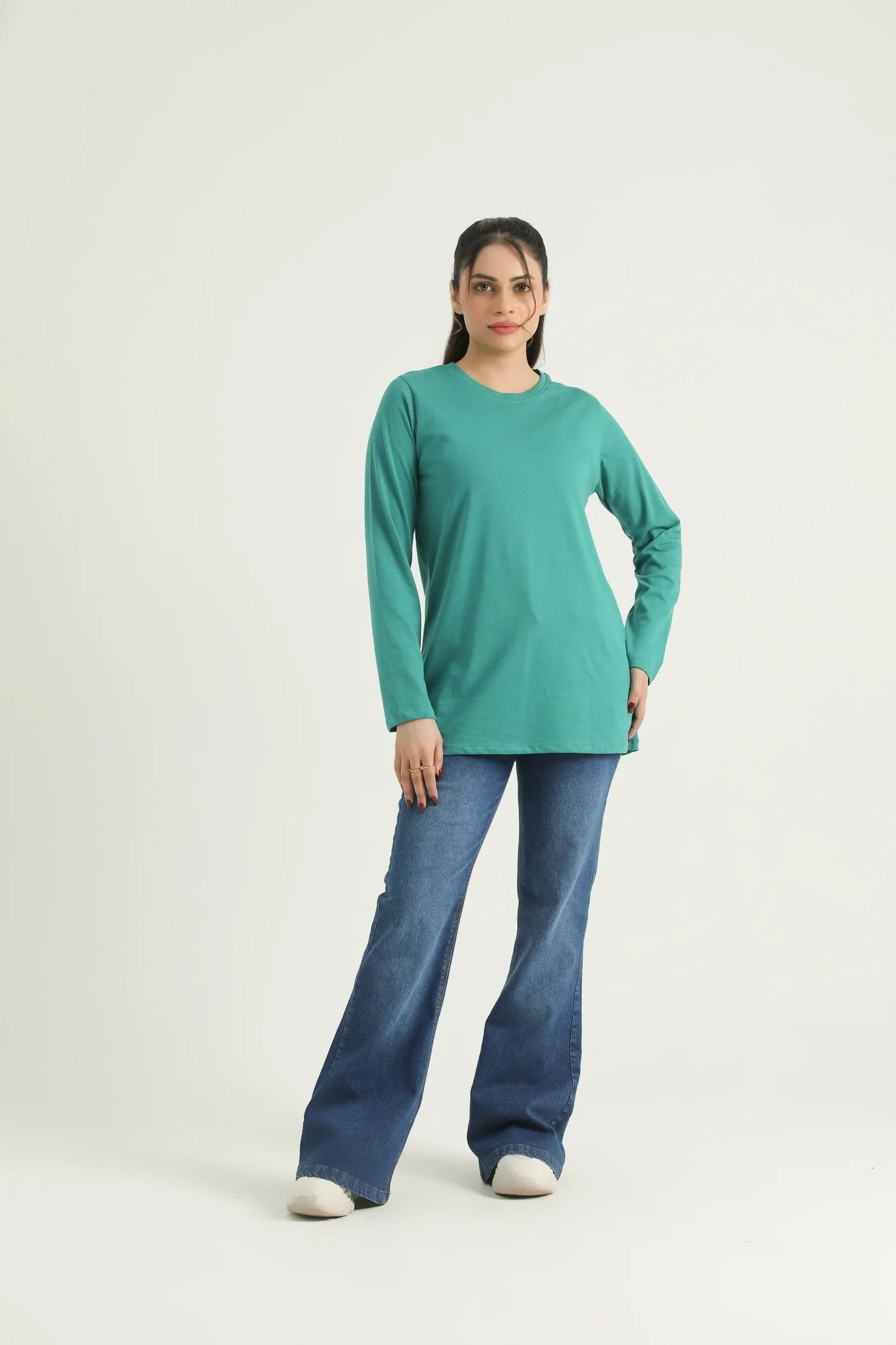 A woman standing against a white background wearing a long sleeve knit top in green and blue jeans.