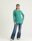A woman standing against a white background wearing a long sleeve knit top in green and blue jeans.