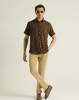 A man wearing a short-sleeved brown linen and cotton shirt with a resort collar and a straight-cut hem with a slit at each side. He is also wearing beige pants and brown shoes.