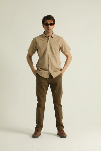 A man standing against a white background wearing a short-sleeved, collarless, beige shirt with a chest pocket, paired with brown trousers and brown shoes.