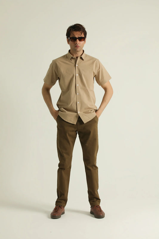 A man standing against a white background wearing a short-sleeved, collarless, beige shirt with a chest pocket, paired with brown trousers and brown shoes.
