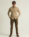 A man standing against a white background wearing a short-sleeved, collarless, beige shirt with a chest pocket, paired with brown trousers and brown shoes.
