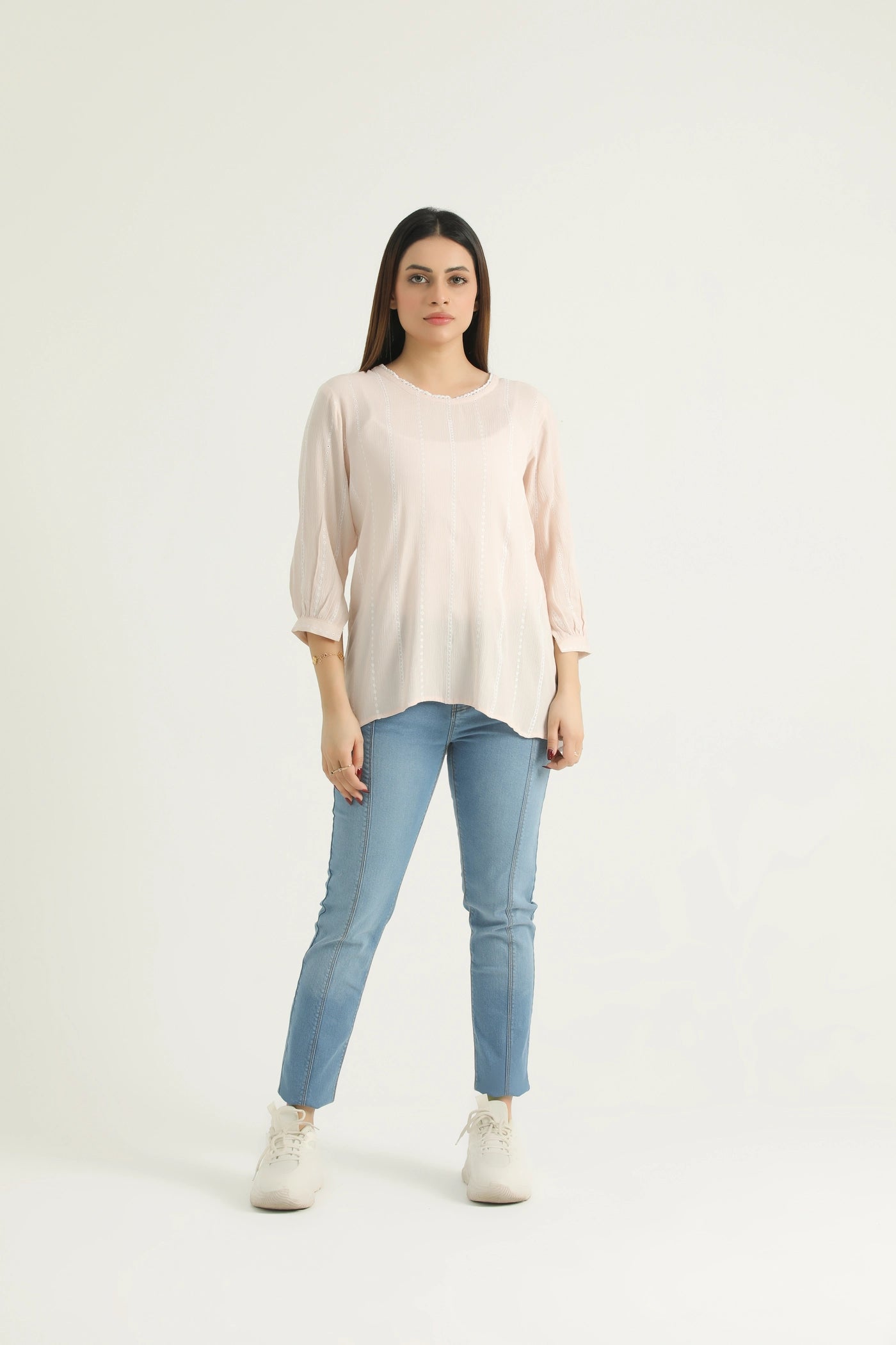A woman standing, wearing a beige tank top with a round neck and back slit opening, paired with blue jeans and white shoes.