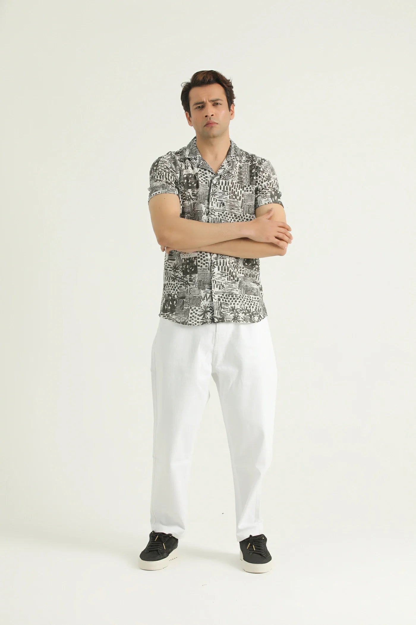 A man standing against a white background, wearing a short-sleeved, textured resort shirt with a resort collar and white trousers.