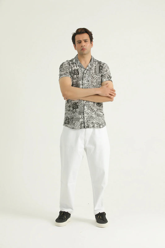 A man standing against a white background, wearing a short-sleeved, textured resort shirt with a resort collar and white trousers.