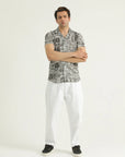 A man standing against a white background, wearing a short-sleeved, textured resort shirt with a resort collar and white trousers.