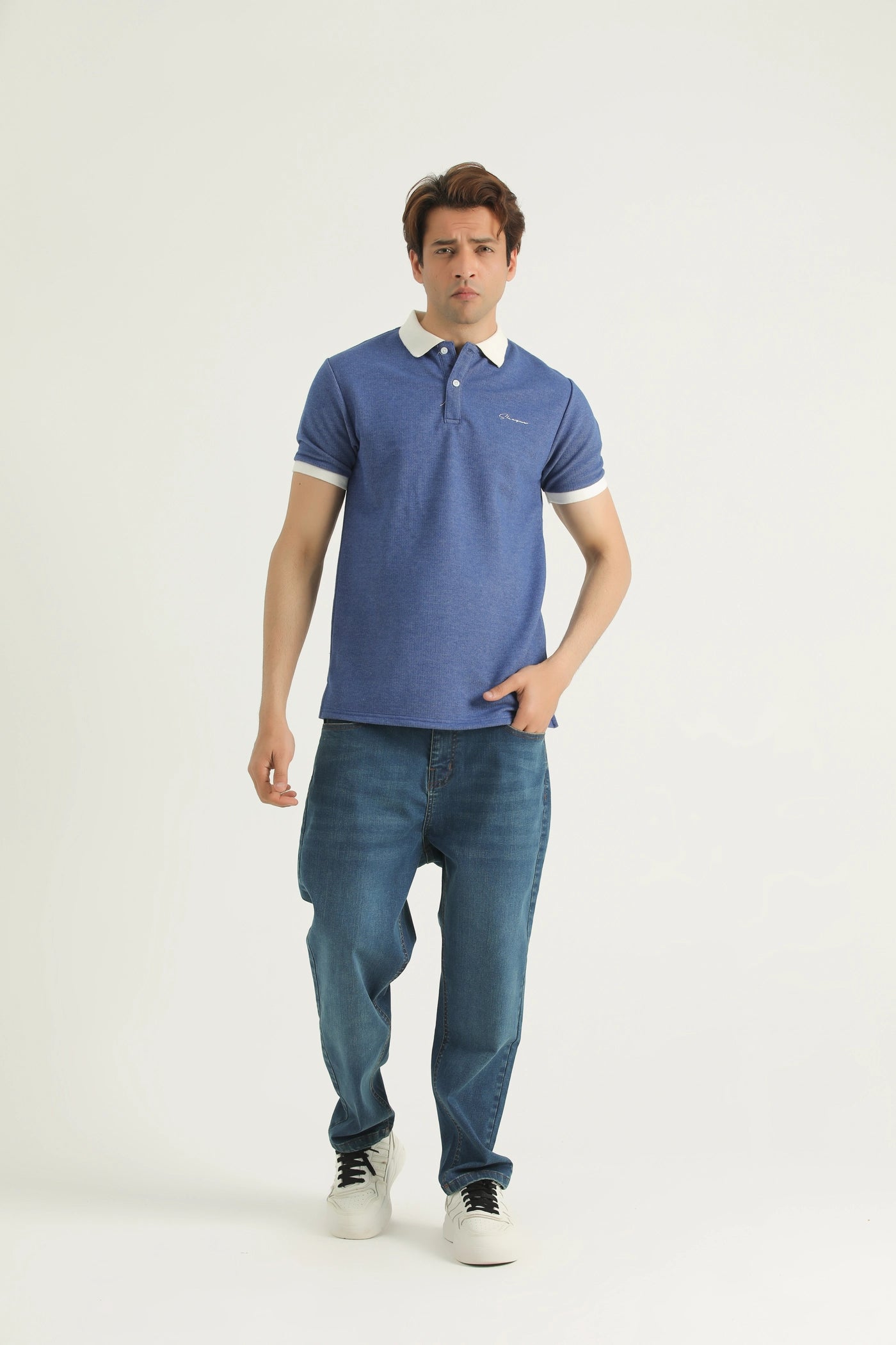 A man wearing a blue textured knitted cotton polo shirt with a collar, short sleeves, and a straight-cut hem with a small slit at each side. He is also wearing blue jeans and white sneakers.